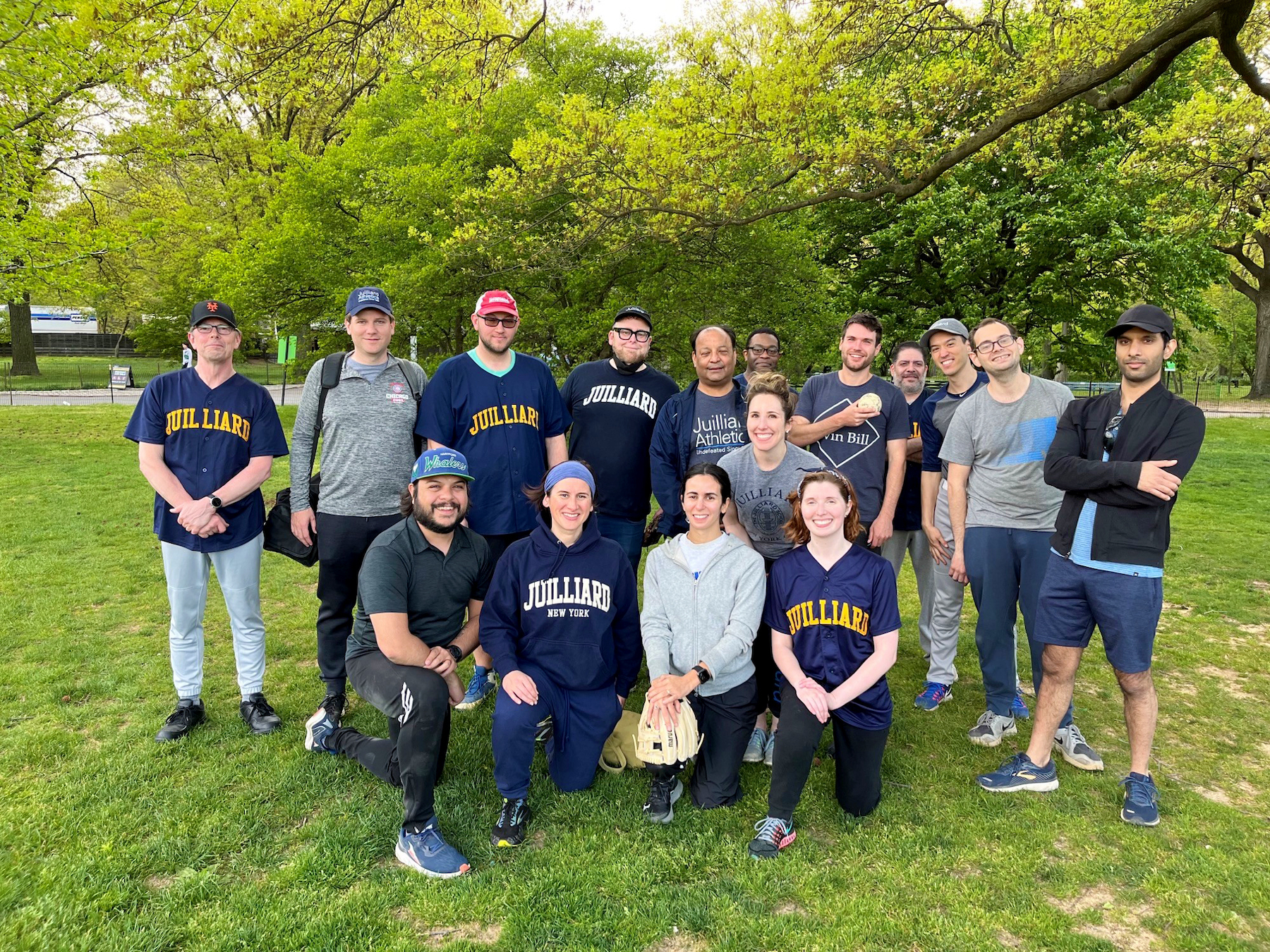 The Juilliard softball team, dressed in their athletic attire, poses in the park, smiling and a sense of camaraderie is present